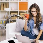women sitting on couch looking t her laptop while drinking coffee