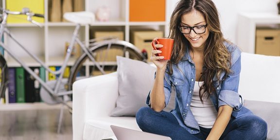 women sitting on couch looking t her laptop while drinking coffee